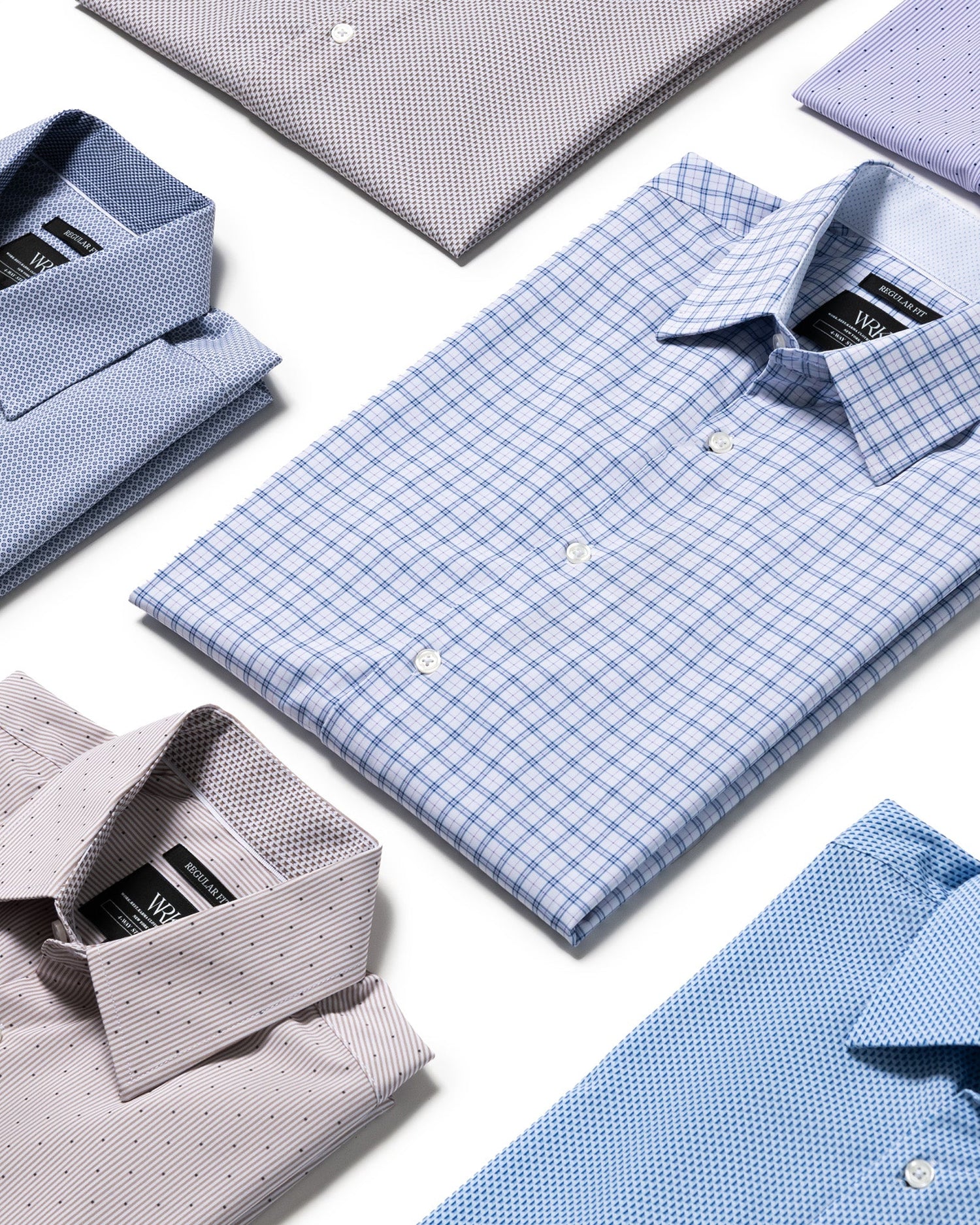 A styled flat-lay of folded dress shirts in various colors and patterns, including a central light blue and white checkered shirt, on a clean white background.